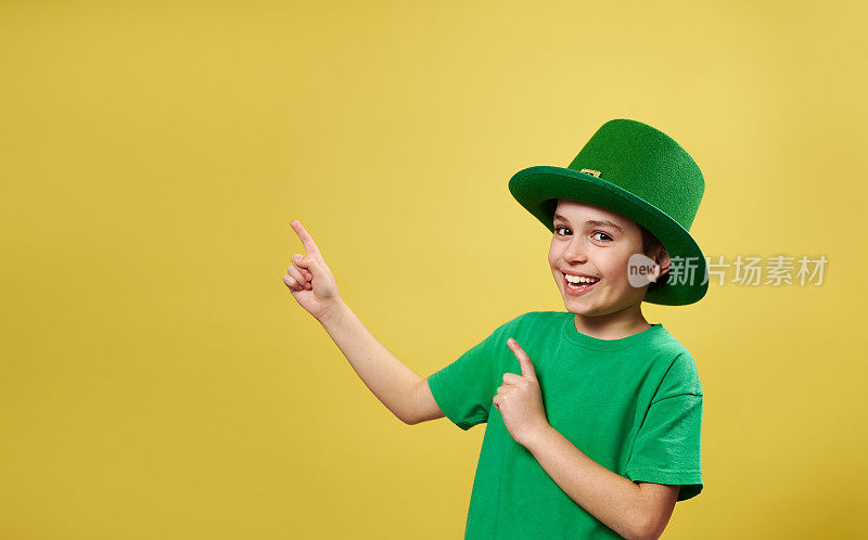 滑稽的小男孩在爱尔兰妖精帽子与他的手指指向和看着相机站在黄色背景与复制空间。圣帕特里克日