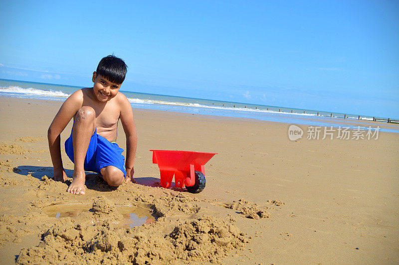 一个快乐微笑的10岁男孩的肖像，穿着蓝色短裤坐在一个红色的手推车在沙滩上的沙子，与大海和海浪和蓝天的背景