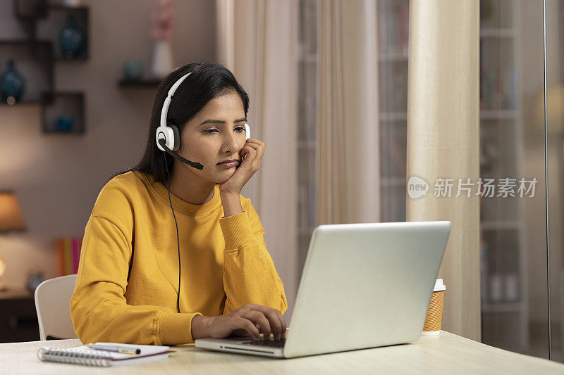 图为一位年轻的女性电话推销员和IT支持人员在家里使用带耳机的笔记本电脑工作