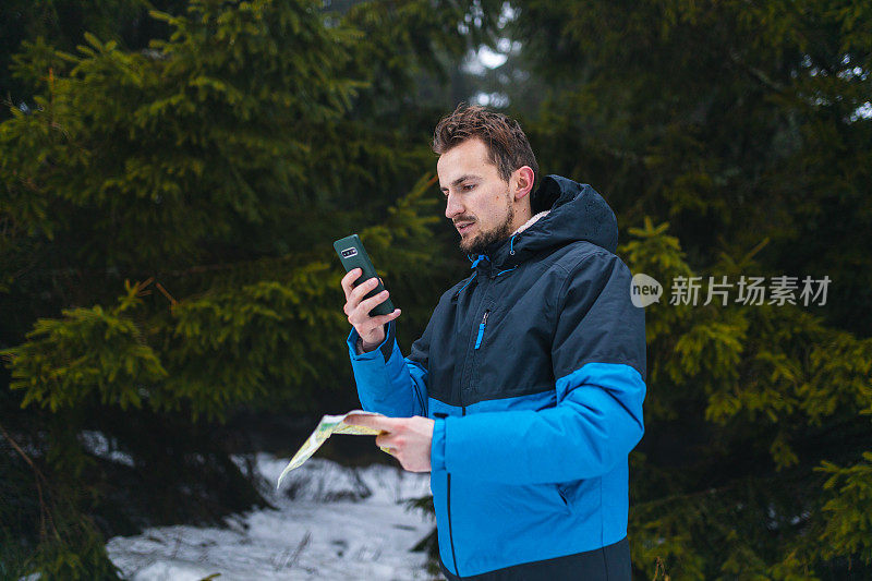 年轻的男性徒步旅行者穿着蓝色夹克，在雾蒙蒙的雪山中迷失了方向，一边看地图，一边看智能手机