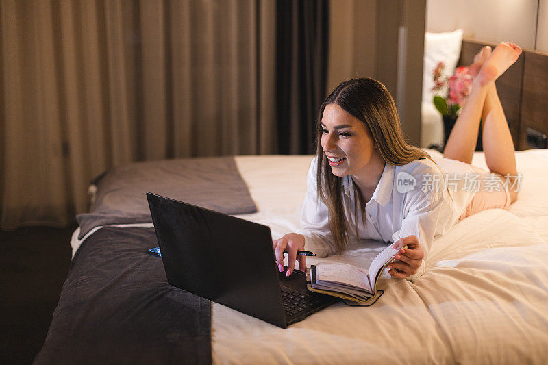 年轻的女商人躺在酒店房间里，一边用笔记本电脑工作，一边在笔记本上记笔记