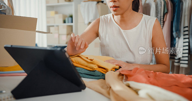 年轻的亚洲小企业主准备包装订单衬衫产品发送客户与库存检查平板电脑在家里。近距离射杀。