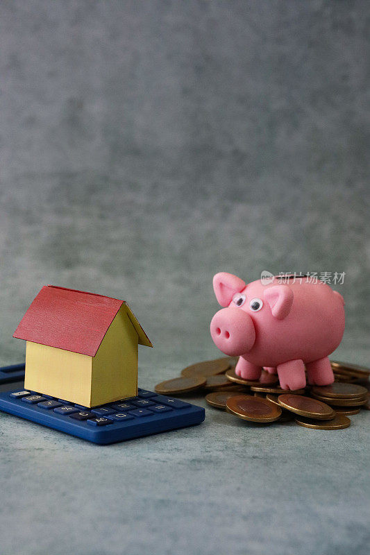 形象的纸板房子站在计算器旁边的小猪银行站在一堆硬币，灰色的背景，抵押贷款，家庭财务和储蓄的概念