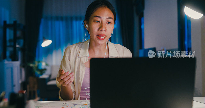 年轻的亚洲女性穿着正装衬衫，戴着耳机，坐在桌子前，笔记本电脑盯着屏幕，在家加班到半夜，在线开会讨论创意。