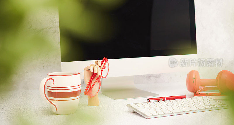 办公室静物配有台式电脑和红色办公用品。咖啡杯和眼镜与叶片模糊在前景