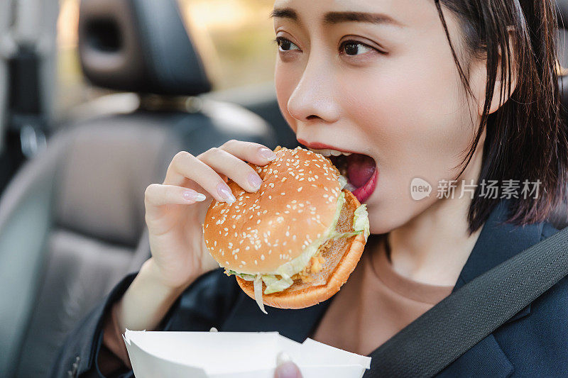 亚洲女人在车里休息，吃汉堡。一个年轻成功快乐的女人正在开车。