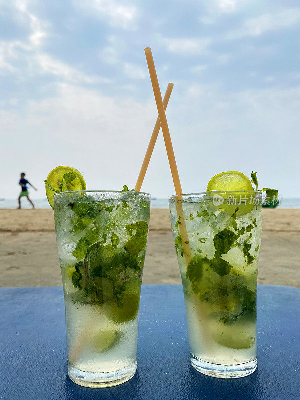 特写图像的海滩餐厅酒吧场景，玻璃杯莫吉托鸡尾酒碎冰，薄荷叶枝，柠檬片和吸管在桌子上，重点在前景