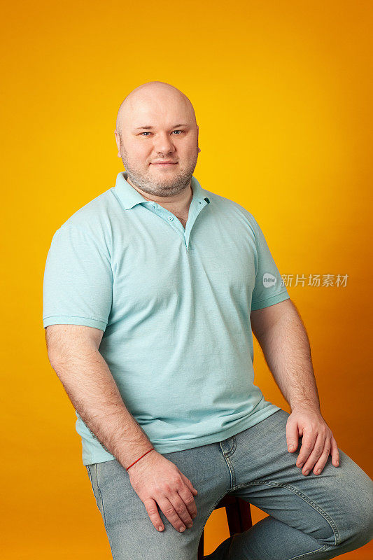 工作室肖像超重中年成年白人男子剃了头，穿着蓝色polo衫，对着黄色的背景