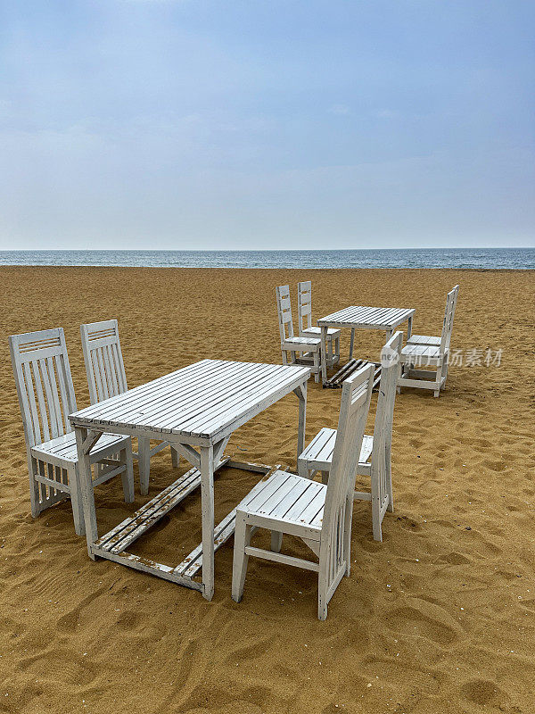 图片中，金色沙滩上质朴的木条餐桌，一组白色的木制桌椅，斯里兰卡科伦坡拉维尼亚山海滩上的游客露天用餐区，蓝色的天空和大海背景，复制空间