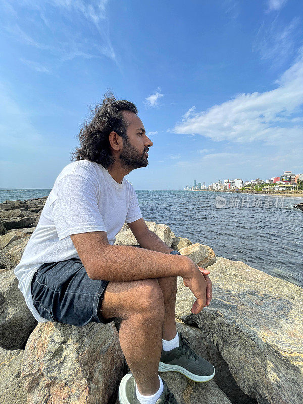 特写图片，印度人坐在斯里兰卡海岸线上的人造岩石海防上，看着风景，海岸侵蚀抛石装甲墙，海岸管理，大海，背景是斯里兰卡科伦坡的城市摩天大楼