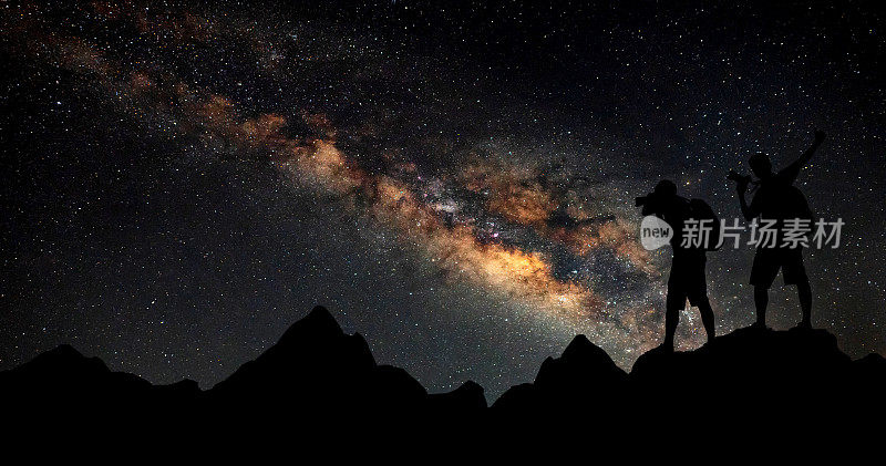 两个摄影师站在岩石上的剪影。暖色夜空与星星和银河与人在山上。高的岩石。背景是星系和一个男人的剪影。许多星星。