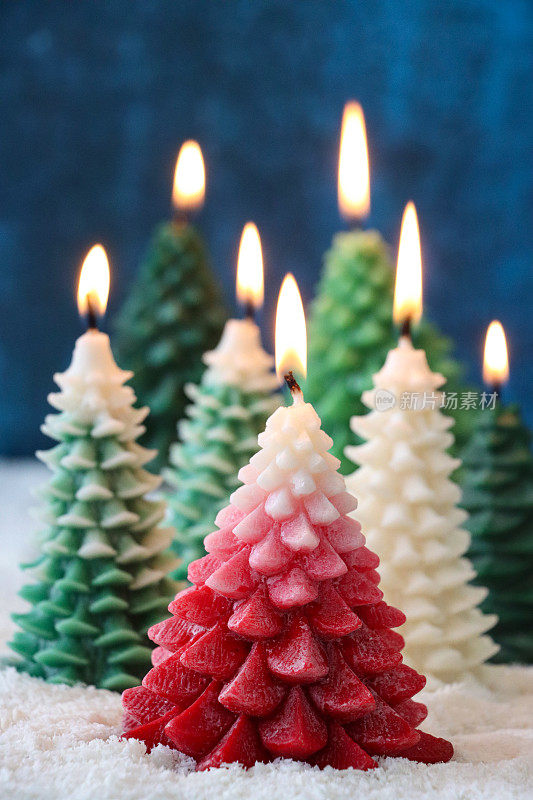 圣诞冷杉森林场景特写图像，一组点燃的圣诞树蜡烛在人造雪上，一棵红色的树被绿色和白色的蜡烛包围，斑驳的蓝色背景，重点在前景