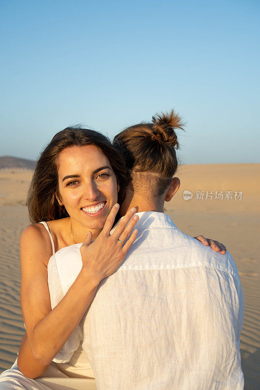 日落时分，一名女子在沙滩上展示订婚戒指，同时拥抱未婚夫