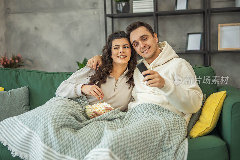 一对夫妇躺在客厅的沙发上，一边看电视一边吃着爆米花