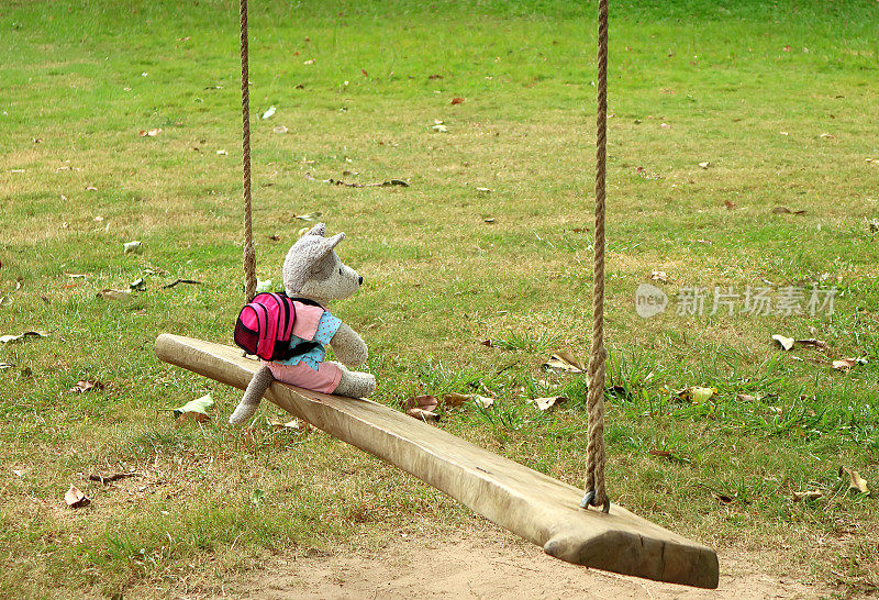 随心!一个可爱的小狗软玩具和背包放松在公园的木制秋千上