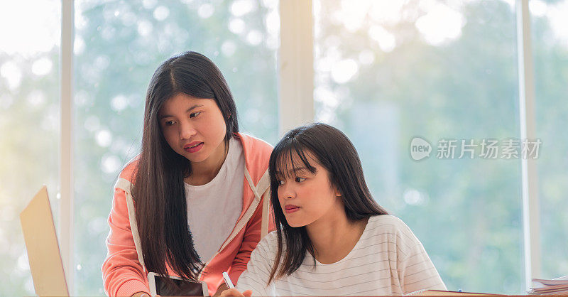 亚洲大学或大学生在图书馆窗口附近与平板电脑、笔记本电脑和报告文件一起学习。快乐的亚洲年轻女子做小组研究在教育概念。