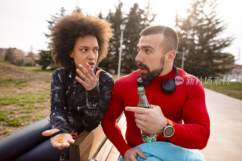 强壮的运动男性和他的伴侣交谈，而她坐在旁边的公园长椅上，手里拿着几颗橙色的药丸