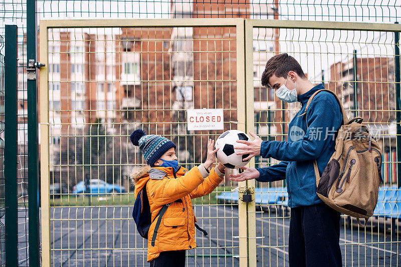 两个男孩在紧闭的操场门前玩球。Covid-19大流行。