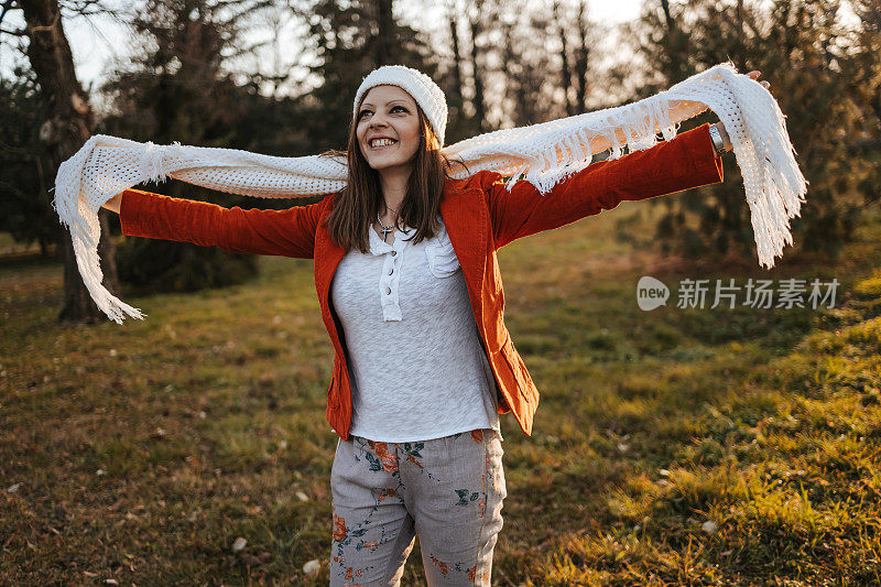 肖像时尚美丽的年轻女子身穿橙色夹克，戴着白色的帽子和围巾，站在秋天的公园里，望着别处