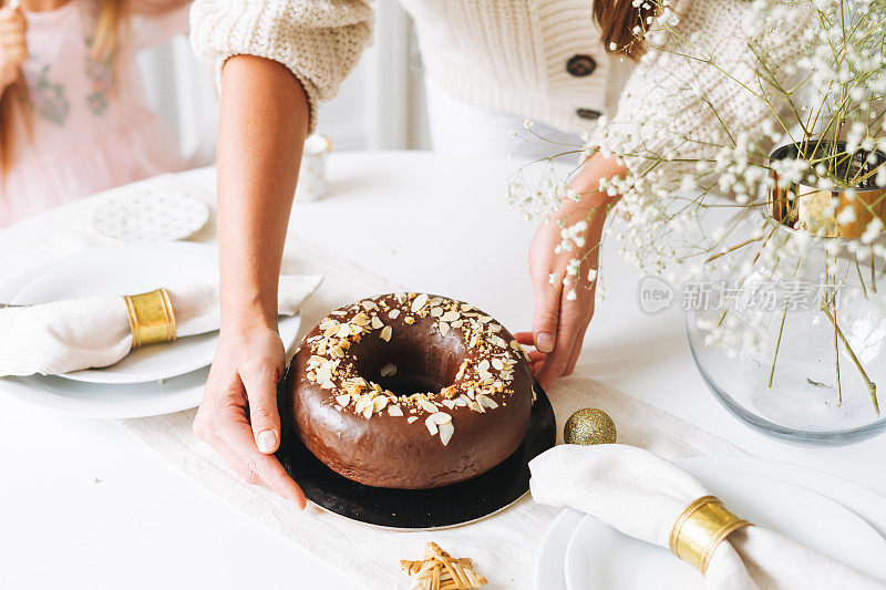年轻的女人母亲手握着巧克力大圆杏仁蛋糕，桌上摆着新年上菜，白色的斯堪的纳维亚餐桌上，家庭大事