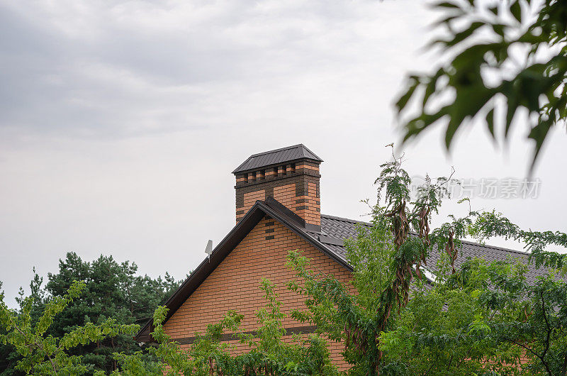 绿树间的小屋。褐色金属瓦砌成的砖房屋顶，窗户、排水沟、潮汐和烟囱映衬着云天。
