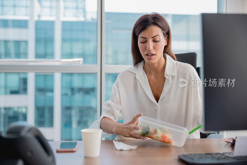 女商人在她的办公室桌子看着蔬菜在塑料盒午餐