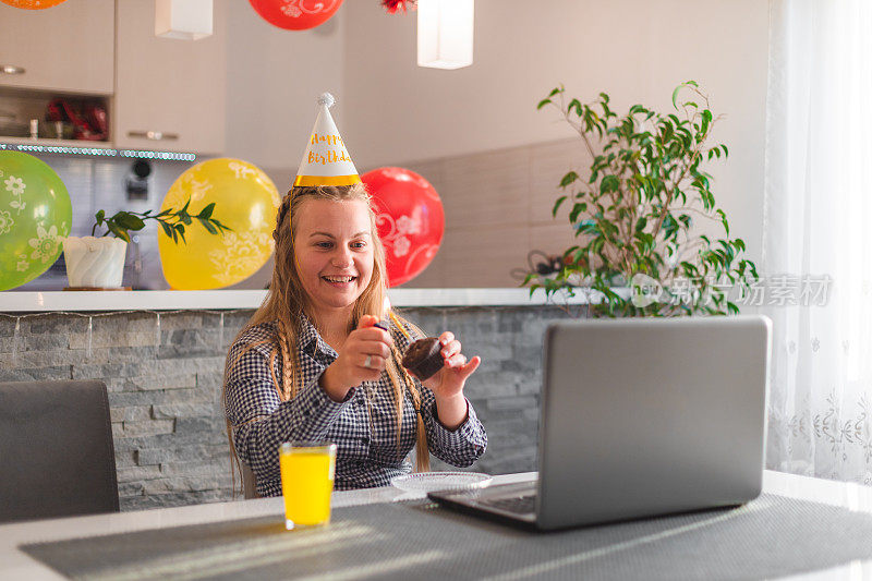 在封锁期间，一名年轻的金发女子在她的笔记本电脑前拿着一个临时制作的生日蛋糕庆祝她的生日