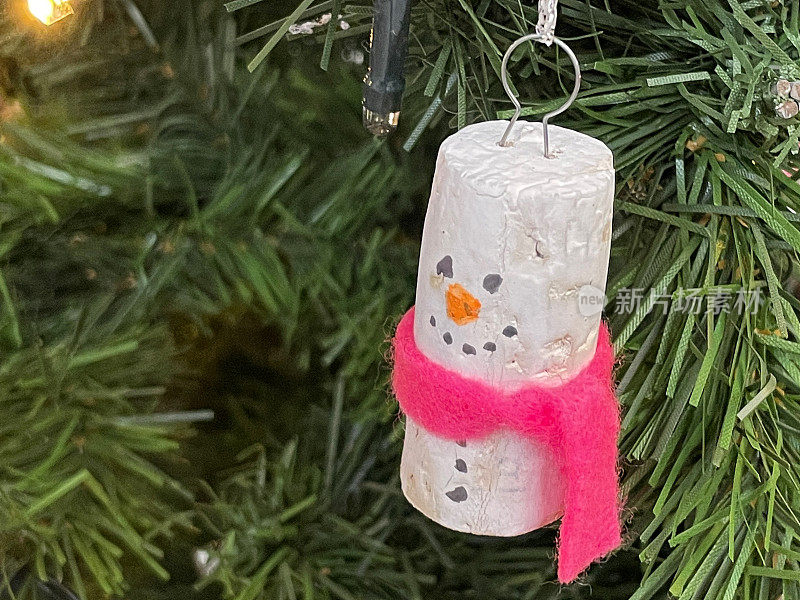 特写图像的软木雪人与粉红色围巾装饰在人造圣诞树树枝，云杉针，模糊的白色精灵灯的背景