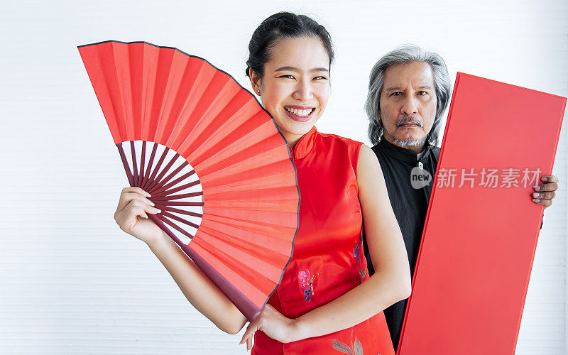 选择性的聚焦美丽幸福的亚洲富家女穿着红色旗袍或连衣裙，微笑着，拿着折扇与模糊的男性保镖，庆祝中国新年。宗教与节日观念。