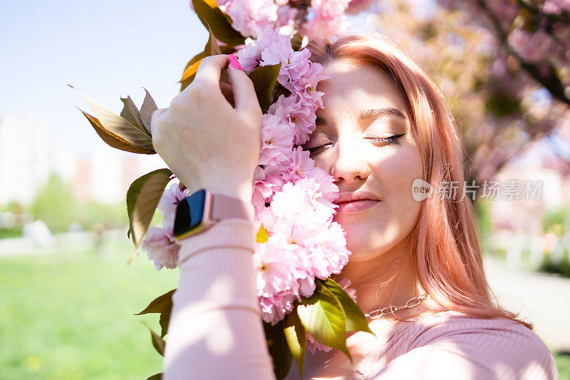 年轻美丽的时尚女孩在一个阳光明媚的日子里戴着帽子摆姿势附近盛开的树与粉红色的花的肖像。春天，女孩在一棵开花的樱花树旁。