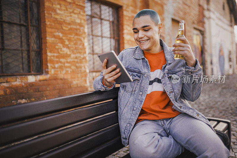 嬉皮士年轻人在公园长椅上用数码平板电脑喝啤酒