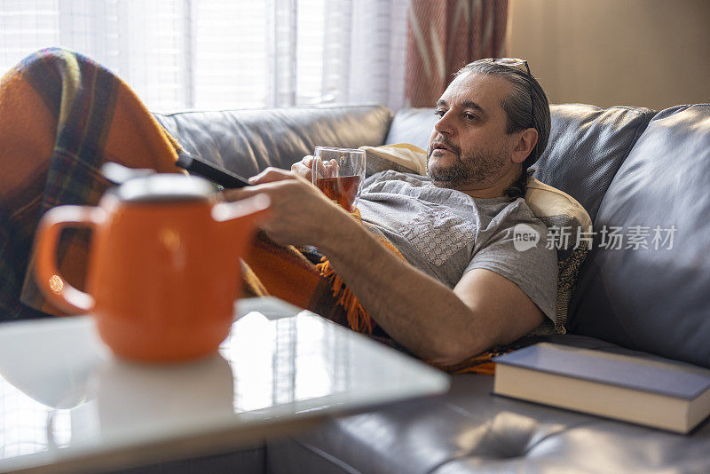 感觉不好。一个成熟的男人生病了，在温暖的格子里躺在沙发上休息。他手里拿着一杯热茶，用遥控器换电视频道。