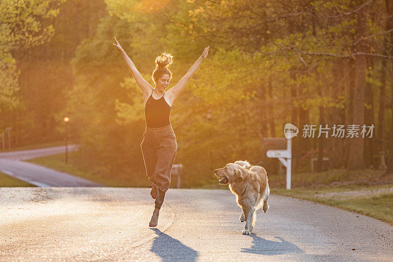 快乐的年轻女子与家庭宠物金毛猎犬在明亮的金色阳光下奔跑