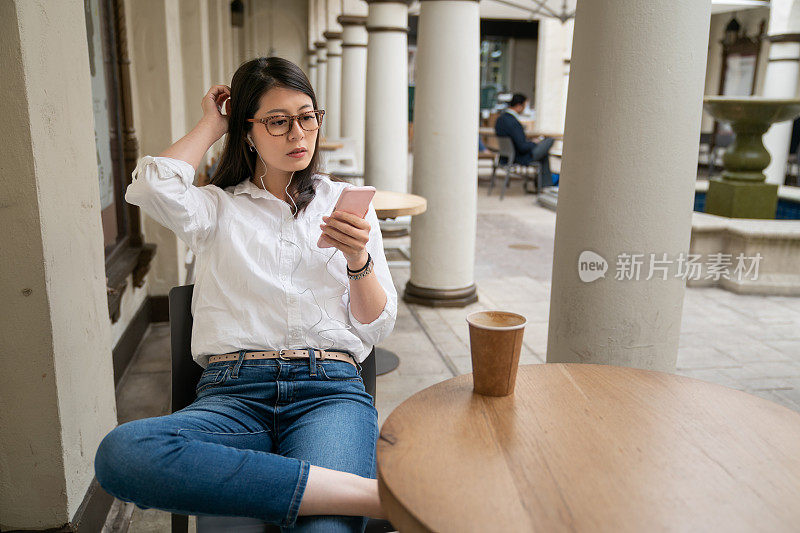美国加利福尼亚州帕洛阿尔托市，亚洲休闲商务女士戴着耳机，午休时间在咖啡馆用手机狂看网络电视节目。她一边看一边折着腿挠着头
