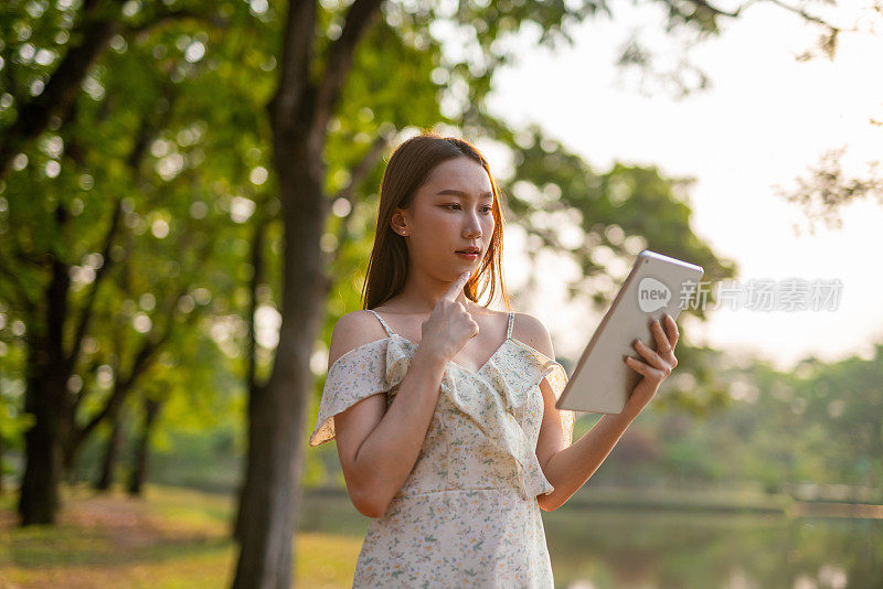 美丽的亚洲女人在公园里玩她的数码平板电脑。在户外工作的年轻女性，一个数字游民