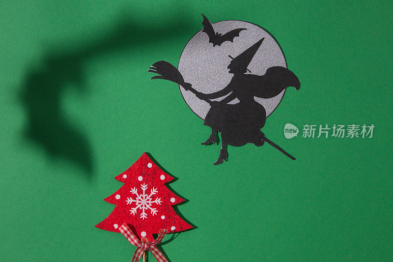 一个女巫骑着扫帚飞到月亮旁边，带着一只蝙蝠和一只蝙蝠的影子飞过圣诞树，在绿色的背景上。复制空间和最小的平面布局。