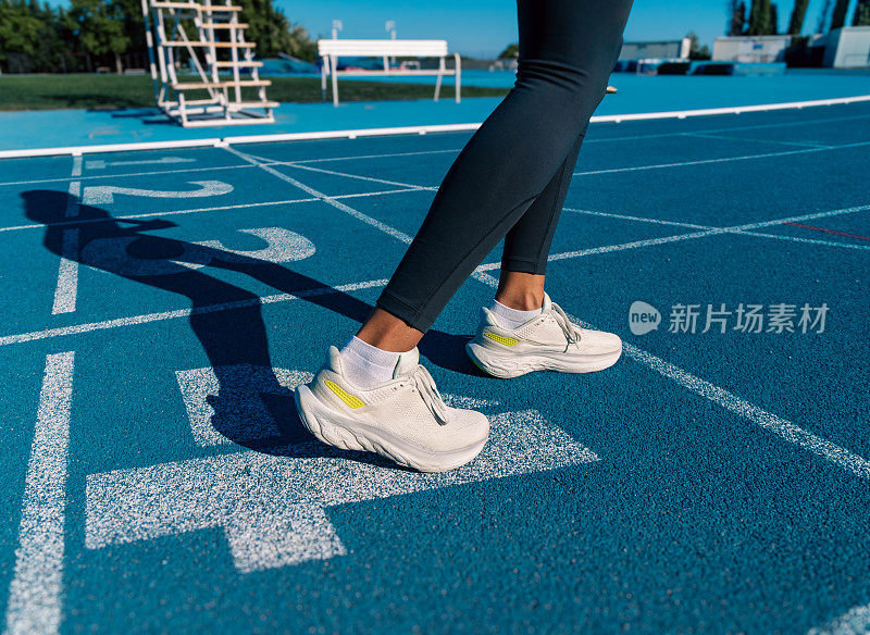 一个年轻的跑步者的腿和运动鞋的影子的细节，准备在一个位置，她的腿和脚弯曲做冲刺或在蓝色田径跑道上比赛。