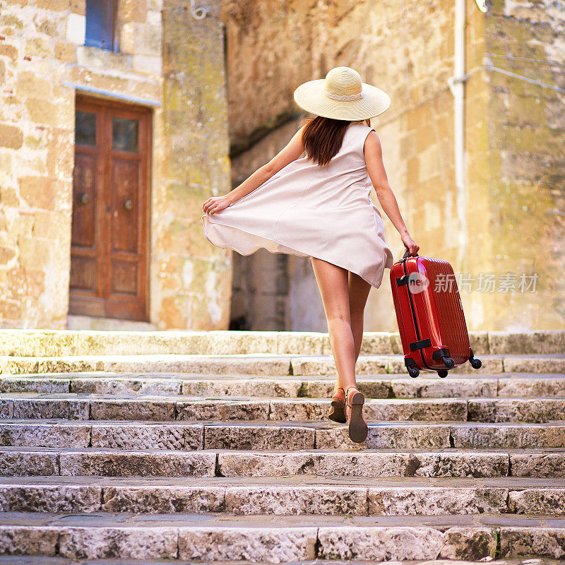一个年轻的女孩，戴着帽子，穿着飘逸的衣服，手里提着一个手提箱，跑上老城的楼梯。从后面看。