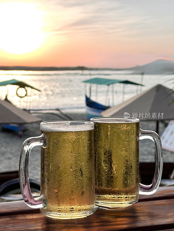 两个玻璃大酒杯里的啤酒，上面布满了凝结的气泡，在啤酒上形成了泡沫，马克杯和品脱玻璃杯放在海滩酒吧户外的木栏杆上，日落时沙滩上的遮阳伞，停泊的渔船，这些都是前景的焦点