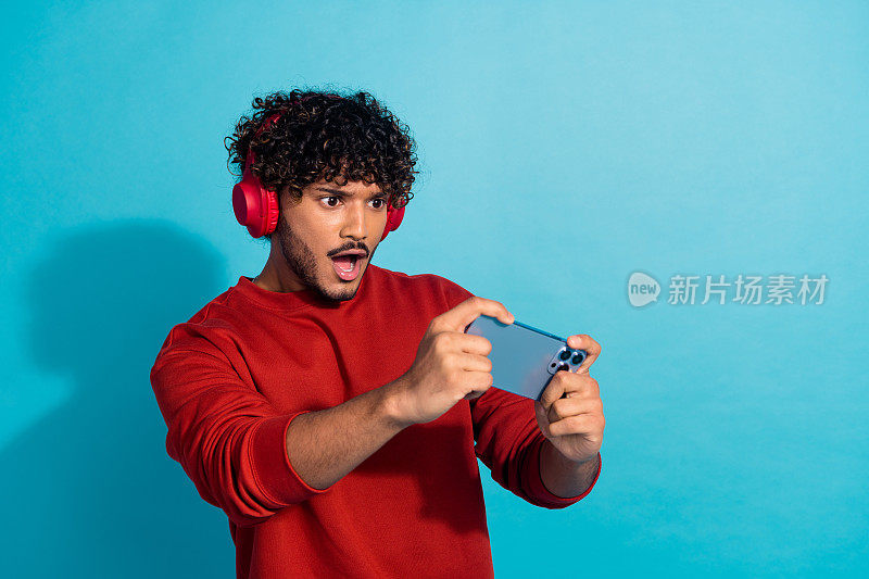 照片的青年有趣的家伙阿拉伯玩手机游戏的智能手机与耳机在机场孤立的海蓝宝石色背景