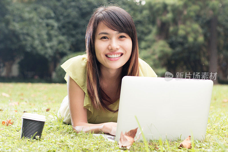 亚洲女学生与笔记本电脑放松在草地上