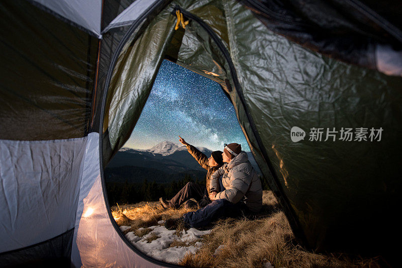 从帐篷里面望去，两位游客正在山里的宿营中休息。男人们坐在篝火旁。一个人指着满是星星和银河的美丽夜空