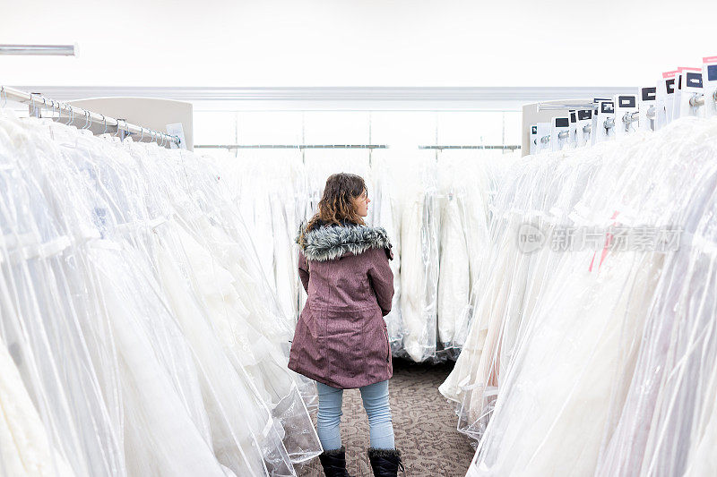 年轻女子在精品折扣店寻找婚纱礼服，许多白色服装挂在衣架排上