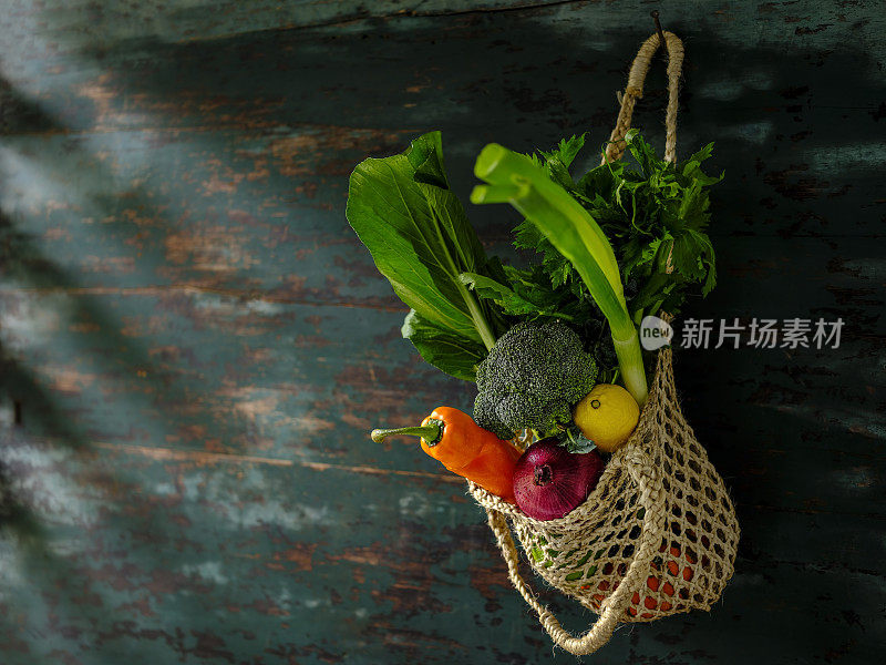 许多色彩鲜明的沙拉蔬菜在一个可重复使用的绳黄麻袋挂在旧风化的木头抽象颜色对比蓝绿色的背景墙。