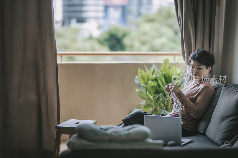一个亚洲华人中年妇女在客厅编织在她的空闲时间，休闲活动和学习从她的笔记本电脑上网
