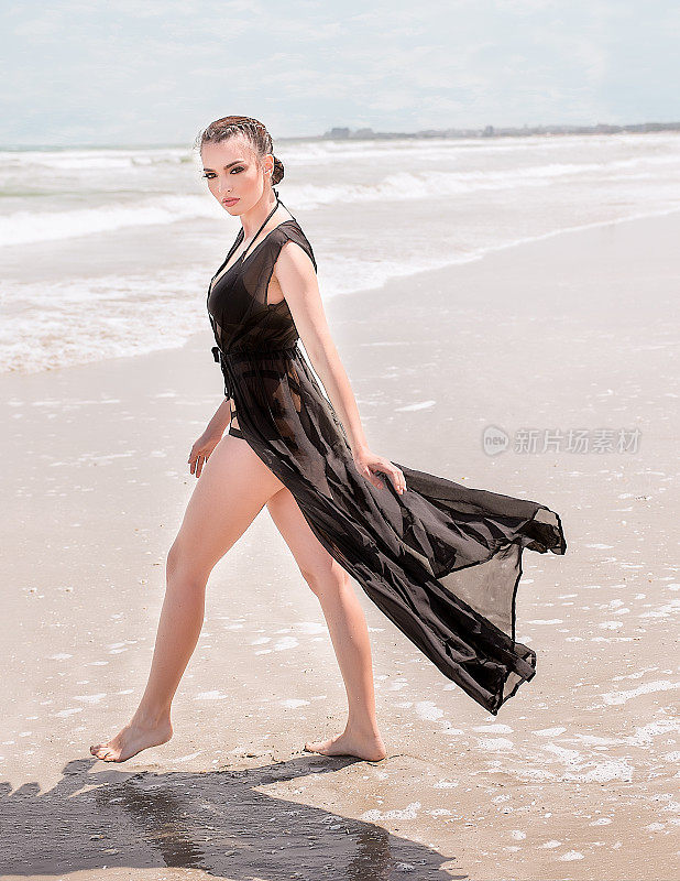 自信的白人女人在侧面，在海滩上与黑色泳衣沙滩透明的裙子和辫子，走到大海