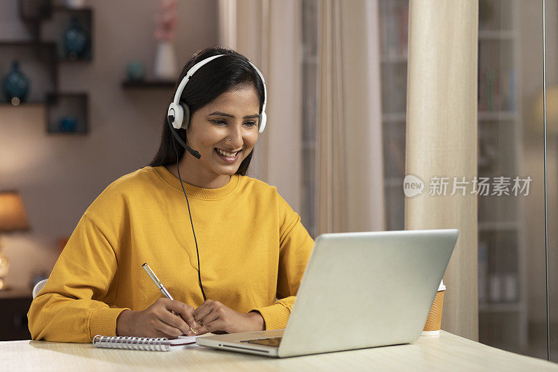 图为一位年轻的女性电话推销员和IT支持人员在家里使用带耳机的笔记本电脑工作