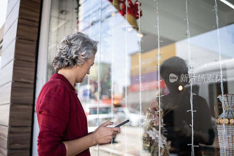 成熟的女人拿着手机看着商店的橱窗