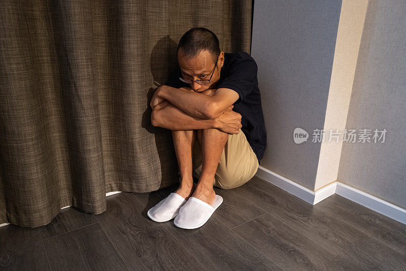 一个亚洲人抱着膝盖坐在房间角落的地板上