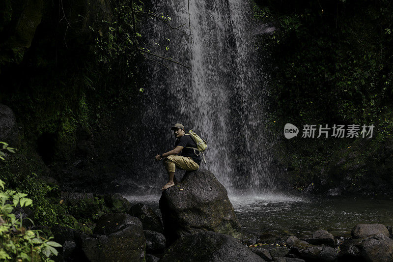 一名亚洲男子走过岩石表面，来到森林中的瀑布处